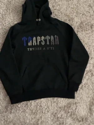 Trapstar svart hoodie med tryck - Svart hoodie från Trapstar med stor huva och metallöljetter. Framsidan har ett färgglatt tryck med texten 'TRAPSTAR' i blått, vitt och grått samt 'IT'S A SECRET' under. Klassisk känguruficka och ribbade muddar. Perfekt för en streetwear-look.