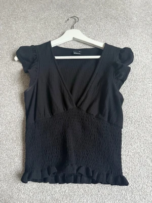 Svart v-ringad topp från Gina Tricot - Säljer en svart topp från Gina Tricot med v-ringning, puffärmar och smockad midja. Toppen har volangdetaljer vid ärmslut och nederkant, vilket ger en söt och trendig look. Perfekt att styla med jeans eller kjol.