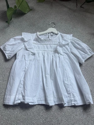 Vit blus med volanger från Gina Tricot - Söt vit blus från Gina Tricot med korta ärmar, volangdetaljer vid axlarna och dekorativ spets över bröstet. Blusen har rund halsringning med liten volangkant och knäppning bak i nacken. Tillverkad i ett lätt och luftigt tyg.