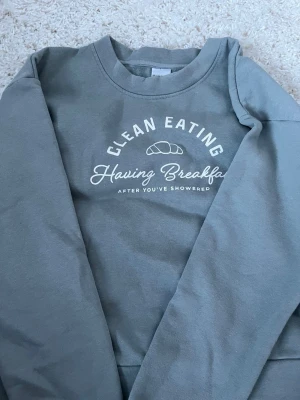 Grå sweatshirt från Kappahl XS - Snygg grå sweatshirt från Kappahl i storlek XS med trycket 'Clean Eating Having Breakfast After You've Showered' och en croissant på bröstet. Tröjan har rund halsringning och är gjord i mjukt, fleeceliknande material på insidan. Perfekt för chill dagar.