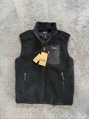 Svart fleeceväst från Patagonia - Säljer en svart fleeceväst från Patagonia med dragkedja framtill, två fickor med dragkedja och en bröstficka i nylon. Västen har hög krage och är gjord i mjukt teddyfleece-material. Perfekt för lager-på-lager och outdoor-vibe. Helt ny och aldrig använd, pris kan diskuteras Storlek Small