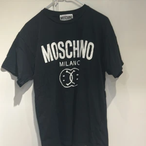 Svart Moschino t-shirt med tryck - Cool svart t-shirt från Moschino med stort vitt logotryck och Milano-text på bröstet. Klassisk passform med rund hals och korta ärmar. Perfekt för dig som gillar streetwear och vill sticka ut med ett ikoniskt märke.
