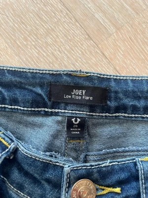 True Religion Joey Low Rise Flare Jeans - Säljer ett par blå True Religion Joey jeans med låg midja och utsvängda ben. Klassisk femficksmodell med snygga kontrastsömmar och flare passform. Använda max 10 gånger 