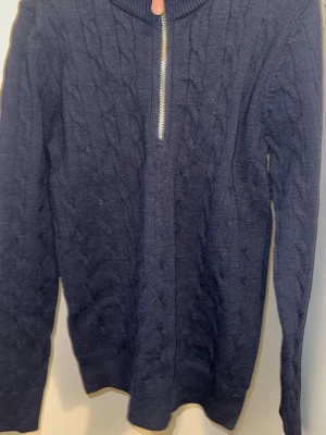 Mörkblå stickad tröja med halv zip – Polo inspirerad (M/L) - Mörkblå stickad tröja med kabelstickat mönster och halv dragkedja framtill. Tröjan har hög krage med en liten läderdetalj vid dragkedjan. Långa ärmar och ribbade muddar vid ärmslut och nederkant. Perfekt för kyliga dagar.