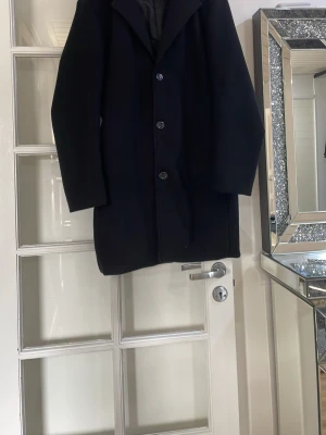 Svart klassisk ullrock - Stilren svart ullrock med knäppning framtill och klassisk krage. Rocken har en rak passform och långa ärmar, perfekt för kyliga dagar. Enkel och tidlös design som funkar till många olika outfits. Inga fläckar, inget smutsigt på den 
