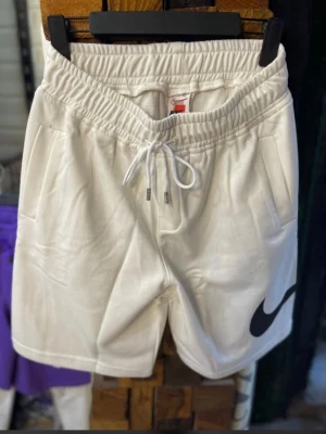 Vita Nike shorts med snörning - Snygga vita shorts från Nike med elastisk midja och snörning framtill. Shortsen har två sidofickor och en stor svart Nike-logga på ena benet. Perfekta för en sportig och avslappnad stil.