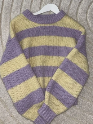 Randig lila och gul stickad tröja - Supermysig stickad tröja med breda ränder i lila och ljusgul. Tröjan har en avslappnad passform, ribbstickade muddar och rund halsringning. 