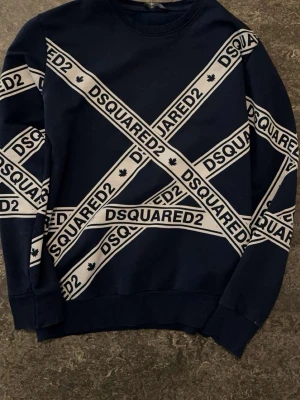 Mörkblå sweatshirt från Dsquared2 - Snygg mörkblå sweatshirt från Dsquared2 med vita diagonala band och logga över hela framsidan. Tröjan har rund halsringning och ribbade muddar vid ärmslut och nederkant. Perfekt för dig som gillar streetwear och vill sticka ut.