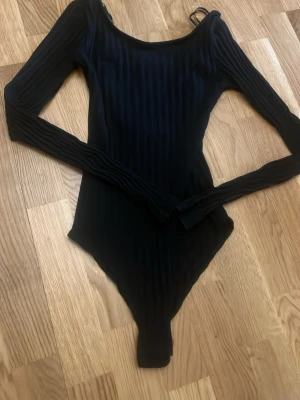 Svart ribbad långärmad body - Snygg svart body med långa ärmar och ribbad struktur. Djup rygg med dekorativa banddetaljer. Stretchigt material som sitter tight och bekvämt. Perfekt att styla med jeans eller kjol för en trendig look.