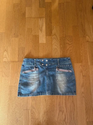 Jeanskjol - Snygg blå jeanskjol i kort modell med två framfickor som har dekorativa dragkedjor. Kjolen har bred linning med dubbla knappar och bälteshällor. Perfekt till en avslappnad och trendig look. Säljer då den inte kommer till användning längre ! Mycket fin med de rosa detaljerna! 💕 storlek s