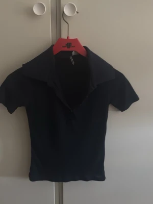 Svart polotopp från Ginatricot - En svart polotopp från Ginatricot med korta ärmar och djup v-ringning. Toppen har en klassisk krage och är figurnära i modellen, perfekt för en stilren look.