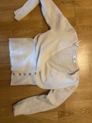 Ljusbeige kofta med knappar - Mysig ljusbeige kofta med v-ringning och fem silvriga knappar framtill. Koftan har lång ärm och är stickad i ett mjukt material som känns skönt mot huden. Perfekt att slänga över en topp för en avslappnad look.