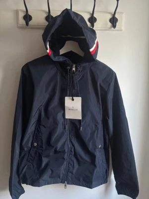 Mörkblå vindjacka från Moncler - Snygg mörkblå vindjacka från Moncler med huva och dragkedja framtill. Jackan har röda och vita detaljer på huvan, två fickor med tryckknapp och elastiska muddar vid ärmslut och midja. Perfekt för blåsiga dagar och enkel att matcha.