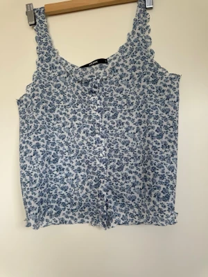 Blommig linnetopp från Bik Bok - Supersöt linnetopp från Bik Bok med blått blommönster på vit botten. Toppen har knappar framtill, volangkant och är ärmlös. Perfekt för varma dagar och enkel att styla med jeans eller kjol.