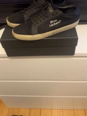 Svarta Saint Laurent sneakers - Svarta sneakers från Saint Laurent med vit sula och handskriven logga på sidan. Klassisk låg modell med svarta skosnören och ovandel i mocka och läder. Perfekta för dig som gillar stilrena och exklusiva sneakers., rätt slitna och släppt från sulan