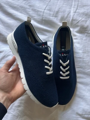 Mörkblå sneakers från Kiton - Snygga mörkblå sneakers från Kiton med stickad ovandel och vita tjocka sulor. Skorna har vita snören och en diskret logga på plösen. Perfekta för en stilren och modern look. Ovandelen är i textil och sulan i gummi.
