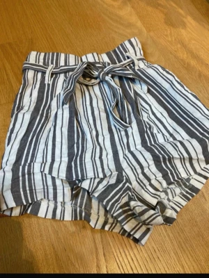 Randiga shorts  - Snygga shorts med grått och vit randigt mönster och knytband i midjan. De har en avslappnad passform och är tillverkade i ett lätt bomullstyg, perfekta för varma dagar. 