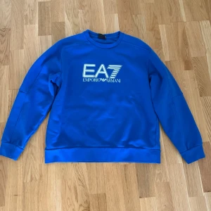EA7 Emporio Armani sweatshirt blå  - Snygg blå sweatshirt från Emporio Armani EA7 med stor logga framtill. Tröjan har rund halsringning, ribbade muddar vid ärmslut och nederkant samt en avslappnad passform. Perfekt för en trendig och sportig look. Pris kan diskuteras 