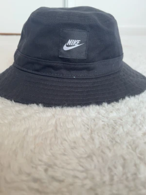 Svart bucket hat från Nike - Säljer en svart bucket hat från Nike med klassisk logga framtill. Hatten är i mjukt tyg och har en avslappnad passform med bred brätte. Perfekt accessoar för en sportig och trendig look.