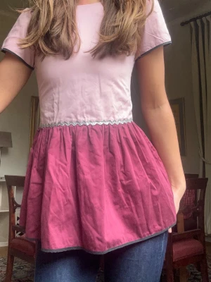 Rosa peplumtop med öppen rygg - ❣️Är från början en klänning som använts som liten från ett lite dyrare barnmärke men inser nu att den kan användas även som topp, ostruken på bilden men såklart finare egentligen❣️Säljer en unik peplumblus i ljusrosa upptill och mörkrosa nedtill. Den har korta ärmar med kontrastkant, markerad midja med volang och en snygg öppen rygg. Materialet känns som bomull och polyester. Perfekt för dig som gillar feminina detaljer. Passar Xs