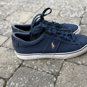 Mörkblå sneakers från Polo Ralph Lauren - Snygga mörkblå sneakers från Polo Ralph Lauren med klassisk låg profil och vit sula. Skorna har canvasmaterial och ikoniska broderade loggan på sidan. Perfekta för en stilren och avslappnad look. Skorna har användsts sparsamt och det förekommer bara lite smuts på sulan som man enkelt kan tvätta bort (se på bilderna). Tveka inte att höra av er vid frågor! 