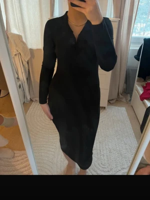 Svart långärmad  klänning tajt  - Säljer en svart, långärmad klänning i ribbat material med figurnära passform och knappar vid halsringningen. Klänningen är midilång och har en enkel, stilren design som passar perfekt till många olika tillfällen.