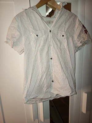 Vit randig kortärmad skjorta Beach Point - Kortärmad skjorta från Beach Point i vit med tunna, diskreta ränder. Skjortan har knappar framtill, två bröstfickor med knapp och broderad röd text på ärmen. Tillverkad i bomull, perfekt för en avslappnad stil.