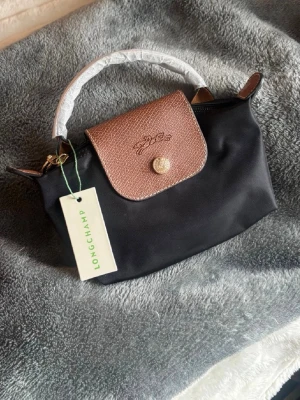 Svart handväska från Longchamp - Snygg svart handväska från Longchamp med brunt lock i präglat läder och guldfärgade detaljer. Väskan har ett kort handtag och dragkedja upptill. Axelrem tillkommer❤️ Helt ny! 