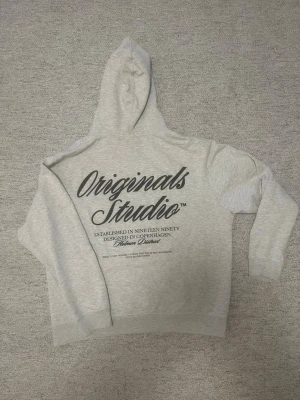 Grå hoodie från Jack & Jones - Snygg ljusgrå hoodie från Jack & Jones Originals med stor svart text på ryggen. Klassisk passform med huva och ribbade muddar. Materialet är mjukt och bekvämt, perfekt för chill dagar. Designad i Köpenhamn med coolt tryck som ger street-vibe.