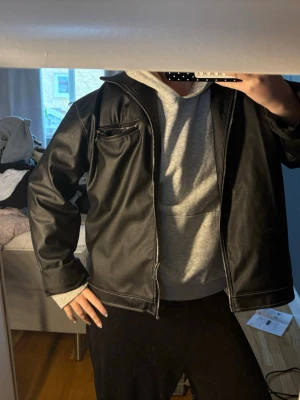 Svart skinnjacka med dragkedja - Snygg svart skinnjacka med silverfärgade dragkedjor och vit kontrastsöm. Jackan har en bröstficka med dragkedja och klassisk krage. Perfekt för dig som vill ha en stilren och tidlös look. Passar till både hoodie och t-shirt. från plt.