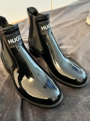 Hugo skor  - Snygga svarta  boots från Hugo i glansigt lackmaterial. Bootsens klassiska siluett kombineras med bred resår på sidorna och tydlig logga framtill. Perfekta för dig som vill ha en stilren men ändå statement look. Materialet är syntetiskt och ger en modern touch. Inga sår eller skador ( kolla bilder ) Luktar inte illa 