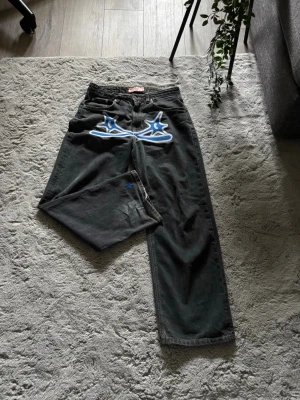 Svarta jeans med blått tryck - Unika svarta jeans med bred passform och stort blått och vitt grafiskt tryck framtill. Jeansen har fem fickor och en liten blå stjärndetalj på ena benet. Perfekta för dig som vill sticka ut med en kreativ och streetinspirerad look.
