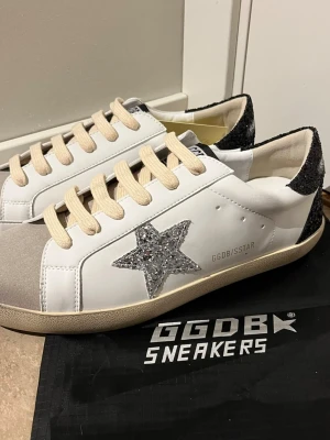 GGDB Superstar vita sneakers med glitter - Snygga vita sneakers från Golden Goose med beige snören, silverglittrig stjärna på sidan och svart glitterdetalj på hälen. Skorna har en beige sula och GGDB/SSTAR-tryck på sidan. Tillverkade i läder med detaljer i textil och glitter.