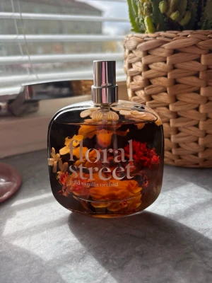 Floral Street Wild Vanilla Orchid parfym - En snygg parfymflaska från Floral Street med doften Wild Vanilla Orchid. Flaskan är genomskinlig med blommigt motiv i orange, rött och gult, och har en silverfärgad spraykork. Perfekt för dig som gillar unika och fräscha dofter. Se bild hur mycket som använts, 50ml flaska. 