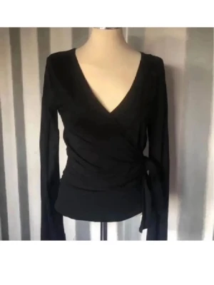 Svart omlottopp med knytband - Snygg svart omlottopp med långa ärmar och knytband i sidan. Djup V-ringning och figurnära passform ger en stilren look. Perfekt att matcha med jeans eller kjol för en trendig outfit.