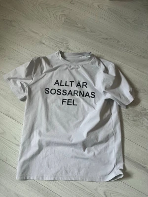 Vit t-shirt med tryck - Vit t-shirt i bomull med svart texttryck på framsidan där det står 'ALLT ÄR SOSSARNAS FEL'. Klassisk rund hals och korta ärmar. Perfekt för dig som vill sticka ut med ett statement-plagg.