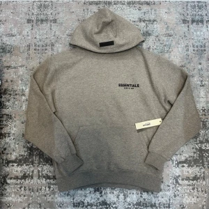 Grå Essentials Fear of God hoodie - Säljer en grå hoodie från Essentials Fear of God med svart logga på bröstet och stor text på ryggen. Hoodien har huva, ribbade muddar och magficka. Tillverkad i mjukt bomullsmaterial, perfekt för en avslappnad och trendig look.