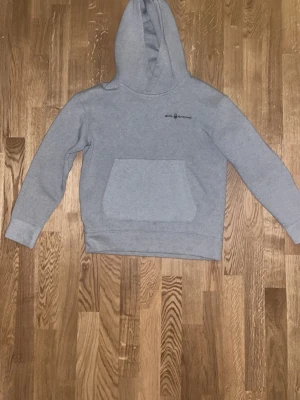 Ljusgrå hoodie från Sail Racing - Ljusgrå hoodie från Sail Racing med klassisk känguruficka och liten logga på bröstet. Hoodien har en stor huva och är gjord i mjukt bomullsmaterial. Perfekt för en avslappnad och sportig stil.