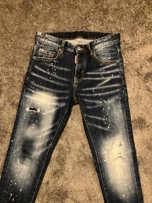 Mörkblå jeans Dsquared2 - Säljer ett par mörkblå jeans från Dsquared2 med slitningar, färgstänk och en skinny fit. Jeansen har klassisk femficksdesign, knappgylf och coola detaljer som ger en trendig streetwear-look. Perfekta för dig som gillar en edgy stil.