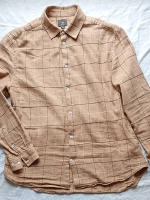 Beige rutig skjorta från H&M - Stilren beige skjorta från H&M med rutigt mönster i svart. Skjortan har klassisk krage, långa ärmar och vita knappar framtill. Tillverkad i ett mjukt, strukturerat tyg som ger en avslappnad look. Perfekt för dig som gillar enkel men trendig stil.