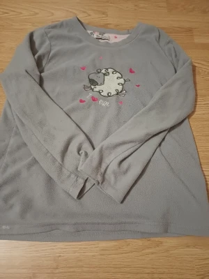 Grå fleece loungewear med fårmotiv - Mysig grå långärmad loungewear-tröja i fleece med ett gulligt fårmotiv och små rosa hjärtan på bröstet. Rund halsringning och avslappnad passform, perfekt för hemmamys. Texten 'ewe' broderad under motivet.