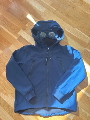 C.P company jacka marinblå - Säljer en mörkblå hoodie från C.P. Company med dragkedja framtill och två snedställda fickor med dragkedja. Huvan har inbyggda glasögon, vilket ger en unik och cool look. Tillverkad i ett mjukt och stretchigt material, perfekt för dig som vill sticka ut.