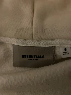 Beige Essentials Fear of God hoodie - Säljer en beige hoodie från Essentials Fear of God i storlek S. Hoodien har en mjuk och fluffig insida, klassisk huva och ribbade muddar. Materialet är en blandning av bomull och polyester vilket gör den både bekväm och slitstark. 