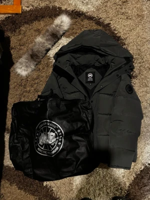 Svart dunjacka från Canada Goose M - Säljer en svart dunjacka från Canada Goose i storlek M. Jackan har huva, avtagbar pälskant och klassisk Canada Goose-logga på ärmen. Flera fickor, dragkedja och knappar framtill. Perfekt för kalla vinterdagar och riktigt snygg streetstil. Ett litet tråd har gått ut från handleden men det märks inte. Undrar du något skriva bara!