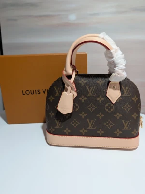 Iouis Vuitton handväska  - Butikspriset är 23 000kr.En elegant handväska i klassiskt Monogram-canvas med detaljer i ljust vachetta-läder. Väskan har den ikoniska strukturerade kupolformen och är utrustad med dubbla handtag samt en matchande clochette (nyckelhållare).