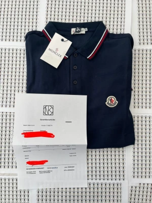 Mörkblå Moncler pikétröja - Snygg mörkblå pikétröja från Moncler med klassisk krage och knäppning framtill. Tröjan har röda och vita ränder på kragen och är dekorerad med Monclers ikoniska logga på bröstet. Perfekt för en stilren och sportig look.