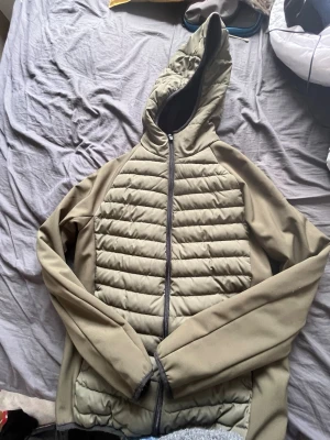 Olivgrön pufferjacka med huva - Säljer en olivgrön pufferjacka med huva och svarta detaljer. Jackan har quiltad framsida och släta ärmar, dragkedja framtill och en sportig look. Perfekt för kalla dagar och enkel att matcha med olika outfits.