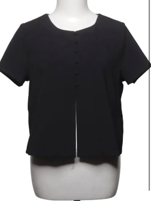 Svart kortärmad blus med knappar - Stilren svart blus med korta ärmar och rund halsringning. Framsidan har en rad klädda knappar och rak passform. Perfekt för en clean och enkel look. Storlek 36 från Gina Tricot
