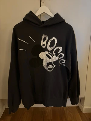 Svart hoodie med Mickey Mouse-tryck - Cool svart hoodie med stor huva och ett grafiskt tryck av Mickey Mouse och texten 'BOO' på framsidan. Oversized passform och ribbade muddar vid ärmar och nederkant. Perfekt för dig som gillar Disney och streetwear vibes.