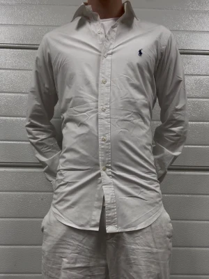 Vit skjorta från Ralph Lauren - Klassisk vit skjorta från Ralph Lauren med knappstängning framtill och broderad logga på bröstet. Skjortan har lång ärm, normal passform och är tillverkad i bomull. Perfekt till en clean och stilren look.